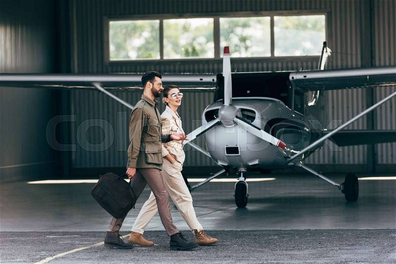 Side view of man carrying bag and walking with stylish girlfriend near hangar with plane , stock photo
