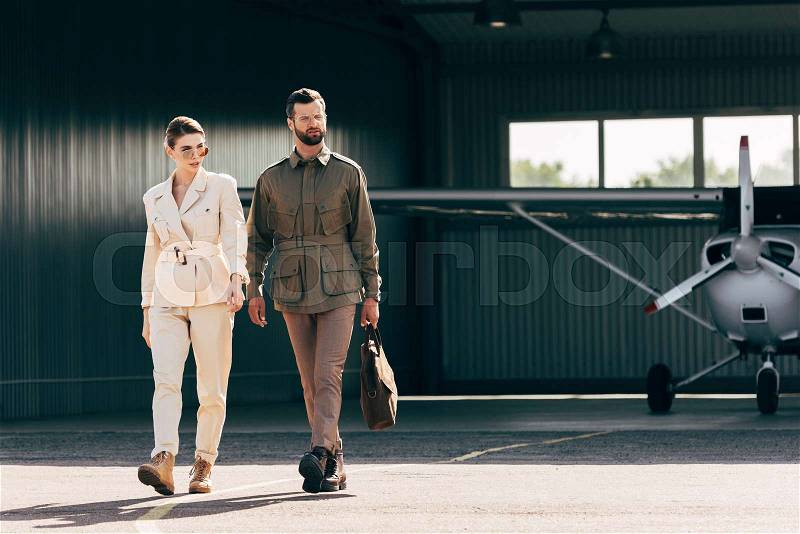 Serious man carrying bag and walking with stylish girlfriend near hangar with plane , stock photo