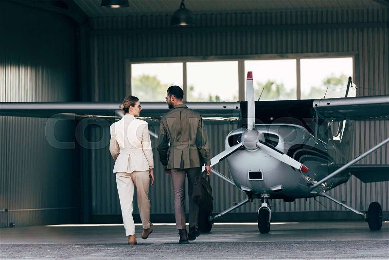 Rear view of man carrying bag and walking with stylish girlfriend near hangar with plane , stock photo