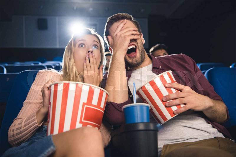 Scared couple with popcorn and soda drink watching film together in cinema, stock photo