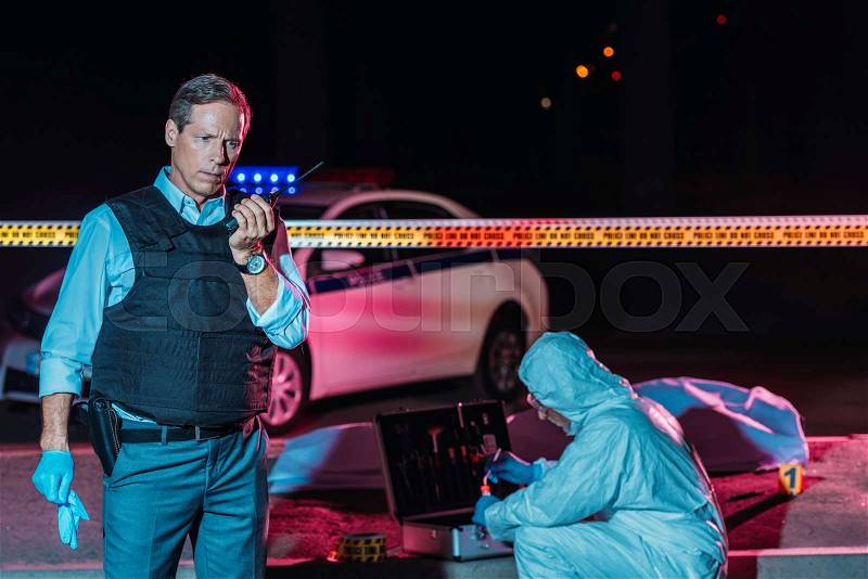 Mature male police officer talking on radio set while criminologist collecting evidence at crime scene with corpse , stock photo