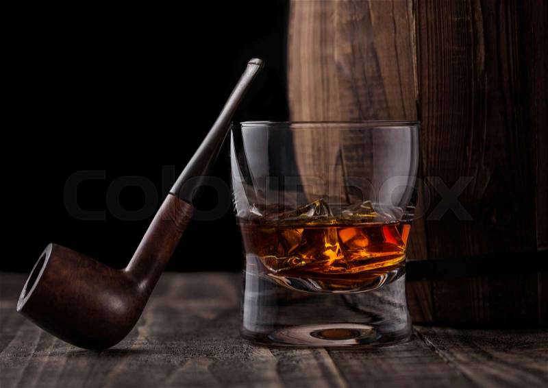 Glass of whiskey with ice cubes and vintage smoking pipe next to wooden barrel. Cognac and brandy drink, stock photo
