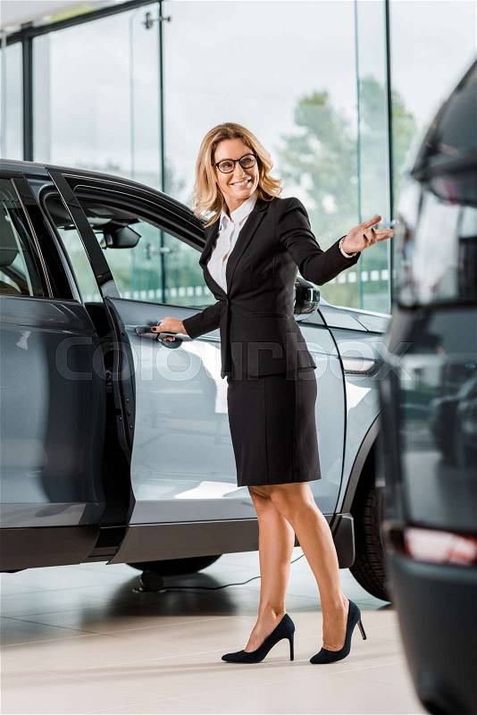Happy female car dealer opening car in showroom, stock photo