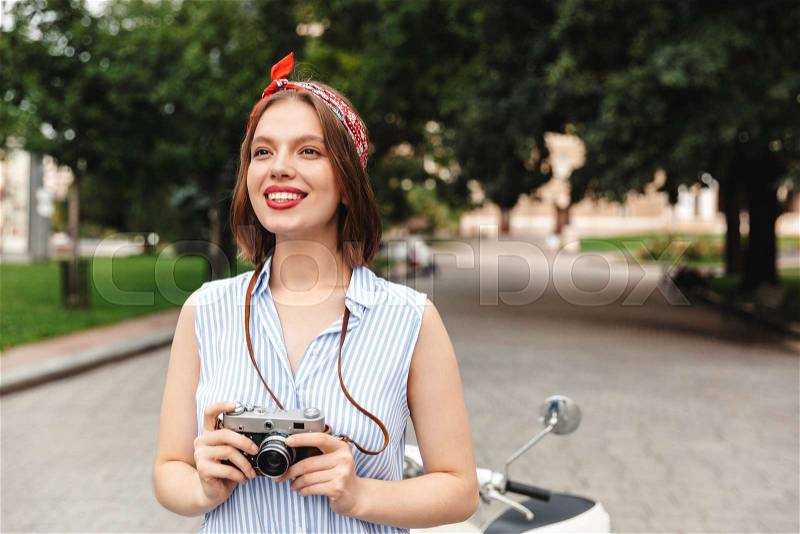 Happy hipster woman holding retro camera and looking away while standing in park, stock photo