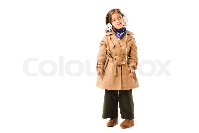 Beautiful little child in trench coat listening music with headphones isolated on white, stock photo