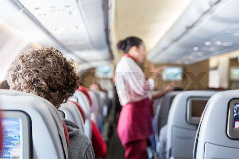 Interior of commercial airplane with unrecognizable passengers on their seats during flight. Stewardess in red uniform at the aisle of commercial airplane, stock photo