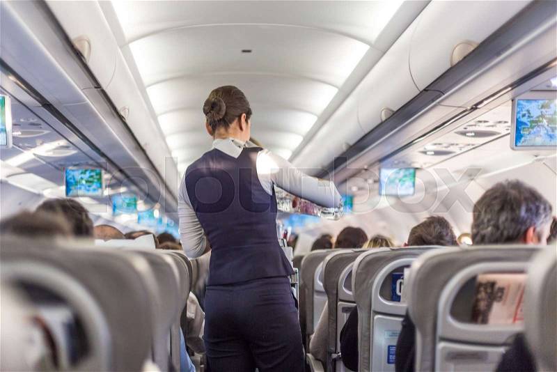 Interior of commercial airplane with flight attandant serving passengers on seats during flight. Stewardess in dark blue uniform walking the aisle. Horizontal ..., stock photo