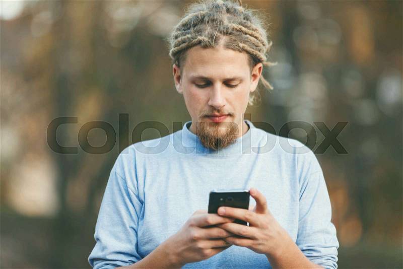 Hipster blonde bearded man with dreads wearing casual clothes watching movie on his smartphone, outdoor view, stock photo