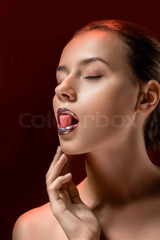 Young woman with shiny lips and closed eyes holding pink candy in mouth on burgundy background, stock photo