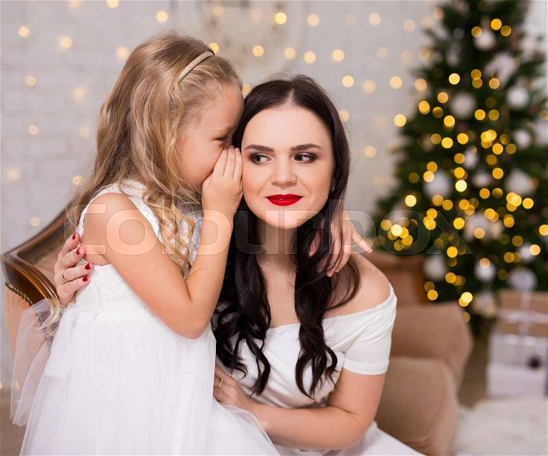 Happy daughter whispering secret or christmas gift wishes to her mother in decorated living room with Christmas tree, stock photo
