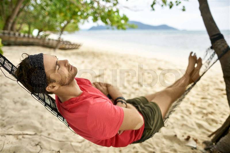 Handsome guy with stubble is lying with closed eyes on the black hammock on the sunny sand beach on the sunny sea background. He wears a red T-shirt, bracelets, dark ..., stock photo