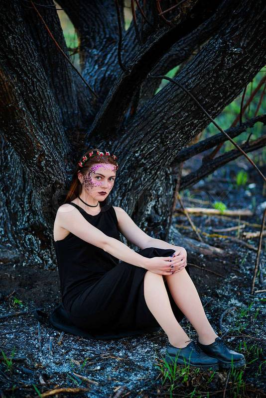 A young beautiful woman with a violet shine on her face stands near a burnt tree, stock photo