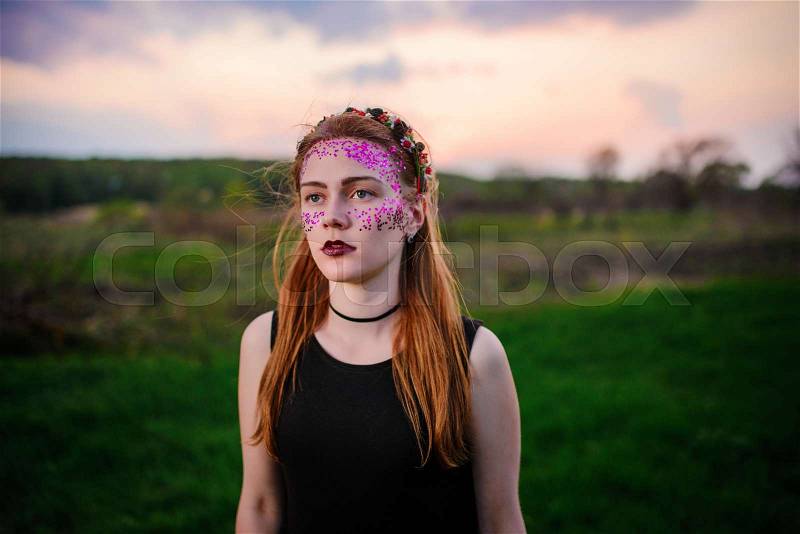 A young beautiful woman with a violet shine on her face standing on the grass and looking at the camera, stock photo