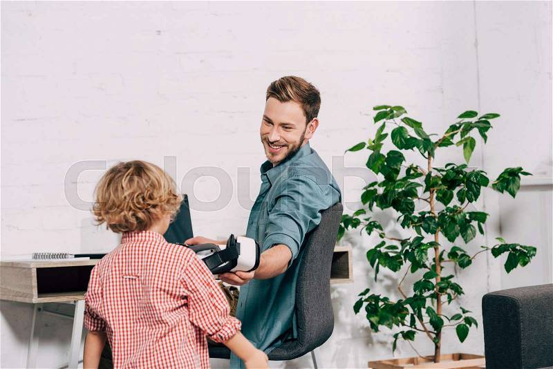 Laughing man giving virtual reality headset to little son , stock photo