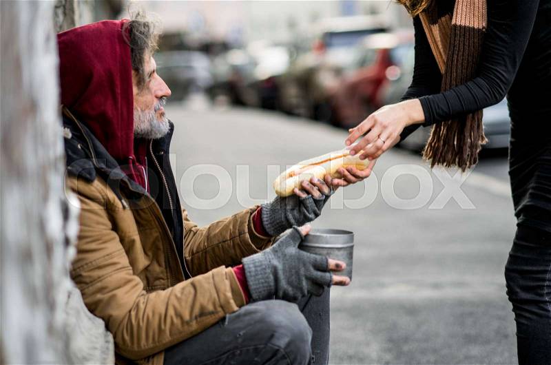 Unrecognizable woman giving food to homeless beggar man sitting outdoors in city, stock photo