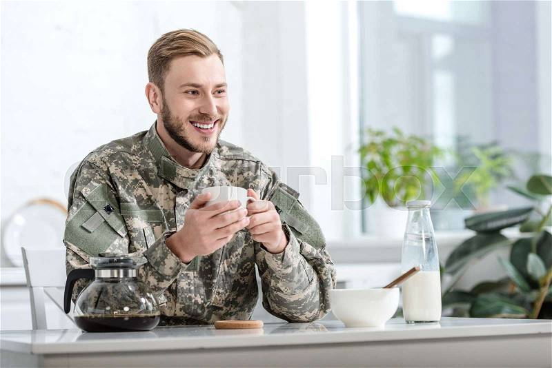 Handsome man in military uniform smiling and drinking coffee at kitchen table, stock photo