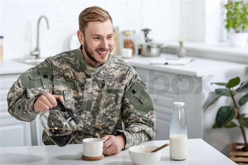 Handsome man in military uniform smiling and pouring coffee in cup from kettle at kitchen table, stock photo