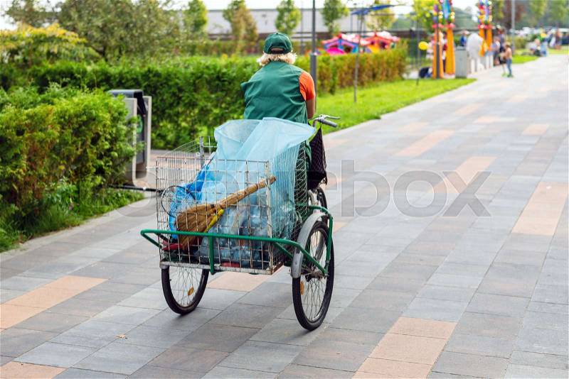 Urban street cleaner riding bicycle with garbage bag through municipal park on sunny day. Sweeper worker with bicycle on pavement at city street, stock photo
