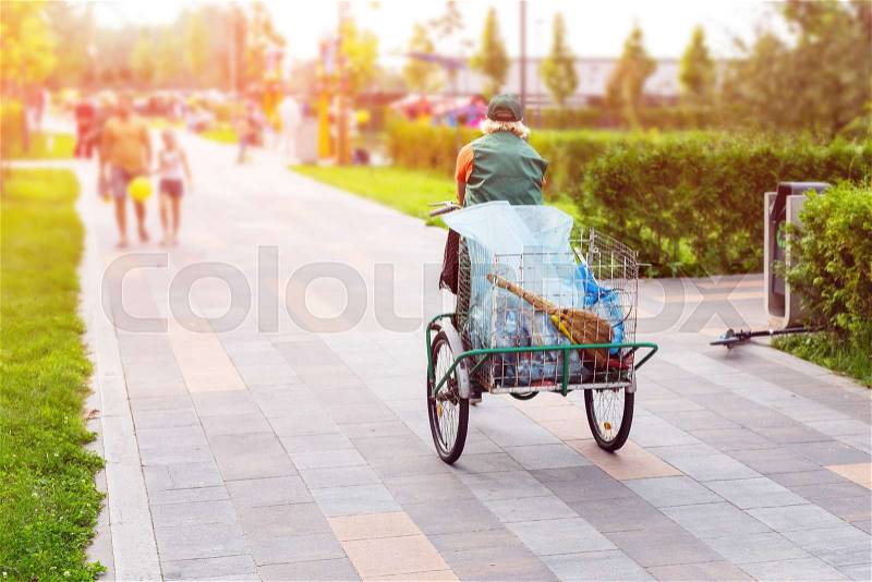 Urban street cleaner riding bicycle with garbage bag through municipal park on sunny day. Sweeper worker with bicycle on pavement at city street, stock photo