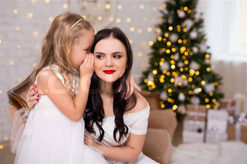 Portrait of happy daughter whispering secret or christmas gift wishes to her mother in decorated living room with christmas tree, stock photo