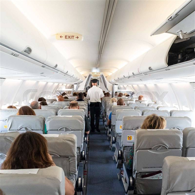 Interior of commercial airplane with unrecognizable passengers on their seats during flight. Steward in blue white uniform walking the aisle of commercial airplane, stock photo