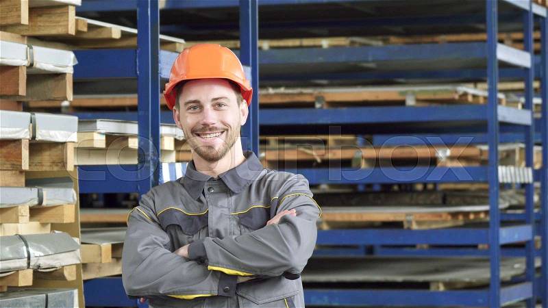 Handsome young male industry worker in uniform smiling joyfully putting on his hardhat standing at the metalworking factory storage copy space profession confidence ..., stock photo