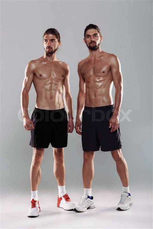 Full length portrait of a two muscular shirtless twin brothers standing isolated over gray background, stock photo