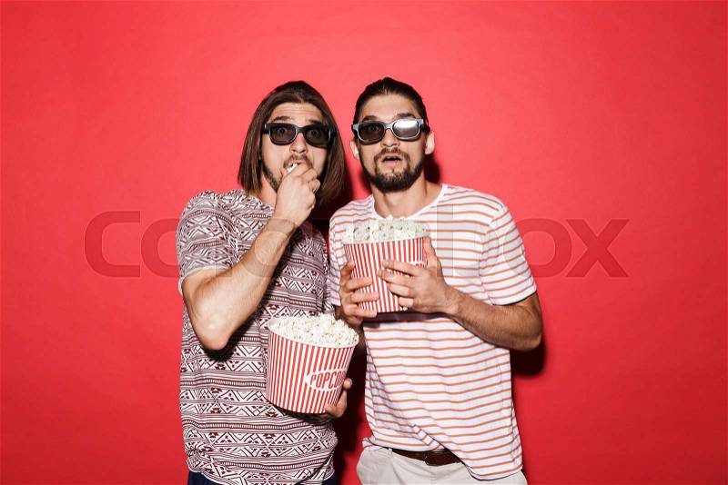 Portrait of a two young frightened twin brothers isolated over red background, watching movie with 3d eyeglasses, eating popcorn, stock photo