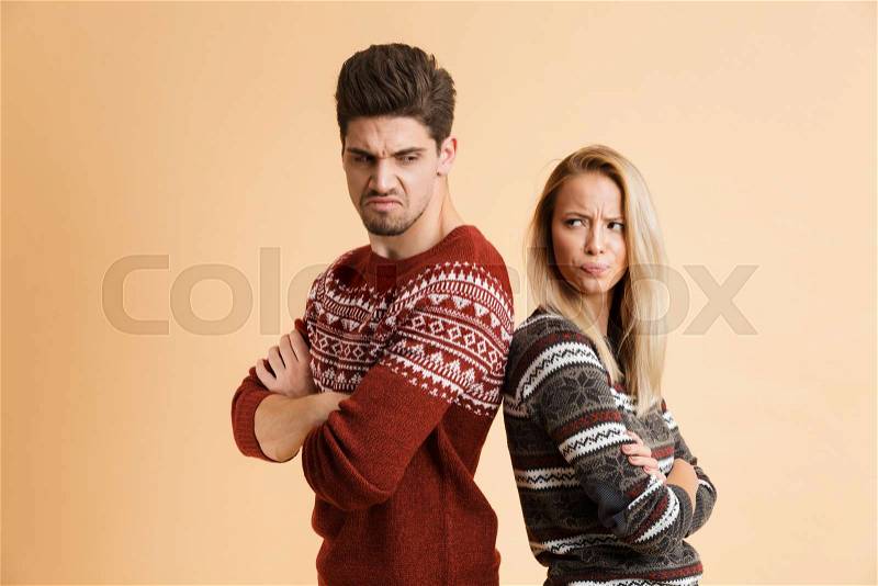Portrait of an angry young couple dressed in sweaters standing together isolated over beige background, arms folded, stock photo