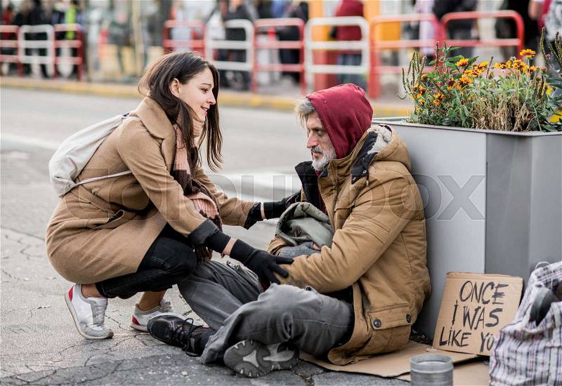 A young woman giving money to homeless beggar man sitting outdoors in city, stock photo