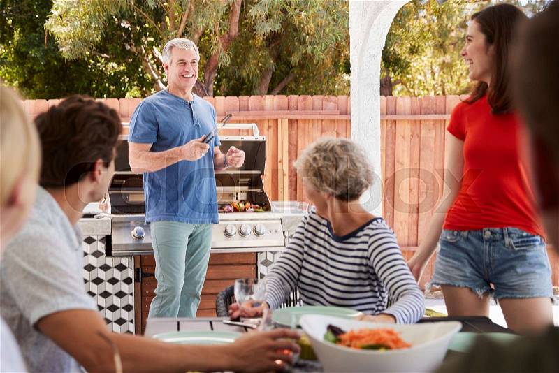 Family at a table outdoors turn to dad standing by barbecue, stock photo