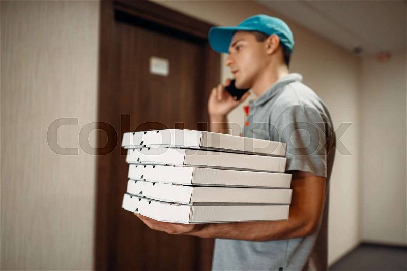 Pizza delivery man at the door calls to customer, delivering service. Courier from pizzeria holds carton boxes and talk by phone indoors, stock photo
