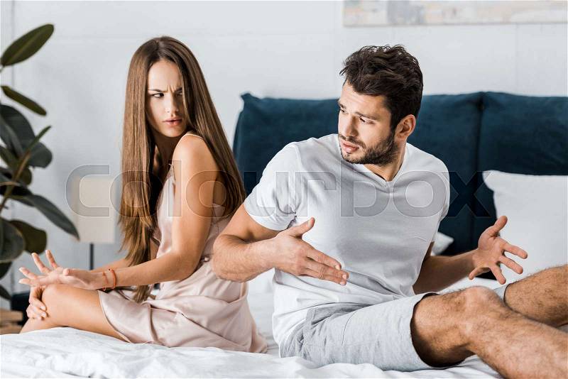 Unhappy frustrated couple in pajamas sitting on bed back to back and gesturing angrily, sexual problems concept, stock photo