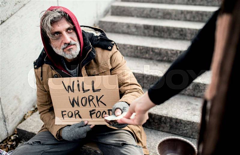 An unrecognizable woman giving money to homeless beggar man sitting in city, stock photo