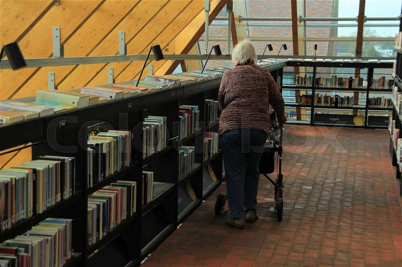 The old lady with her rollator is walking along the collection of books in the municipal library, Boekenberg in the city Spijkenisse, stock photo