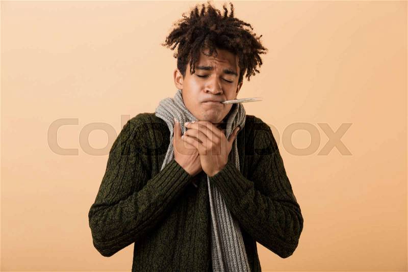 Portrait of frozen african american guy wearing sweater and scarf grabbing throat with thermometer in his mouth, isolated over beige background, stock photo