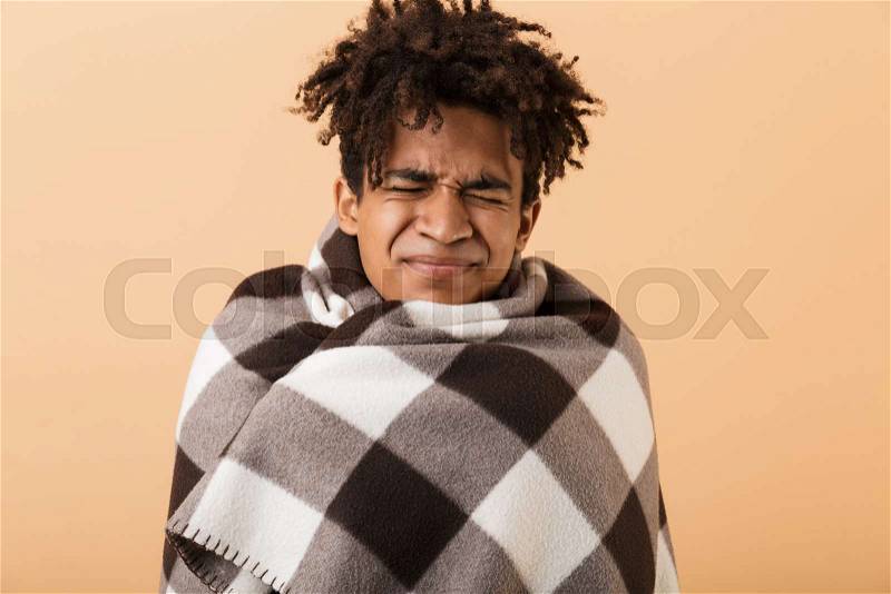 Portrait of frozen african american guy frowning while covering in blanket, isolated over beige background, stock photo