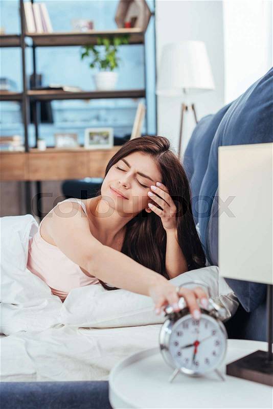 Sleepy girl turning off alarm clock while laying on bed during morning time at home, stock photo