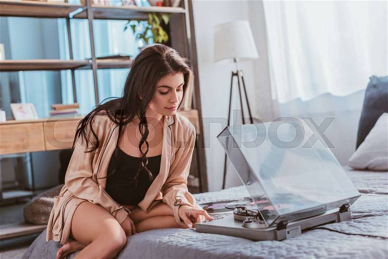 Beautiful girl sitting on bed and turning on vinyl audio player at home, stock photo