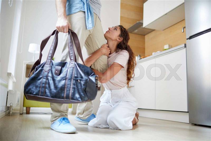 Young knelt woman apologizing and holding her husband with bag by his leg while begging him to stay, stock photo