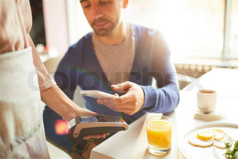 Close-up of handsome man paying bill through smartphone and putting device to cash terminal held by waitress in cafe, stock photo