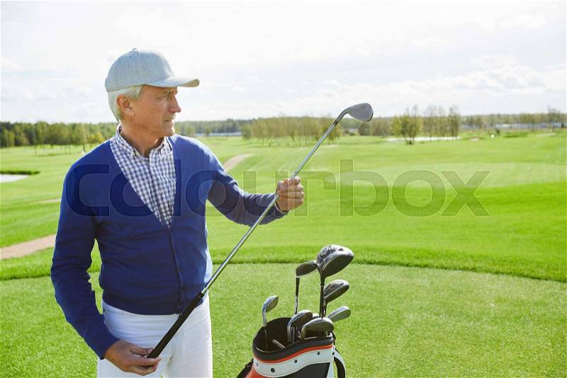 Mature golf player choosing suitable club while preparing for outdoor game on green field, stock photo