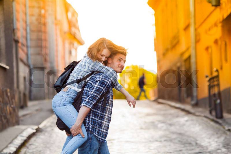 Young man carrying adorable woman on his back while walking on the streets with historical buildings. Old buildings in the rays of golden sun on the background. ..., stock photo