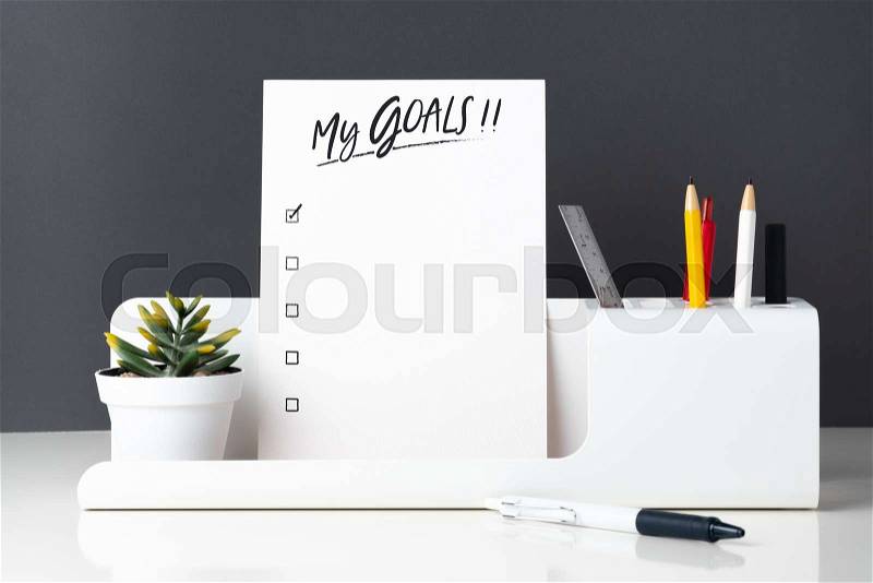 My goals list on notepad at modern office stationery on white table and dark grey wall.pencil, notepad,ruler,pen and pencil box with plant, stock photo