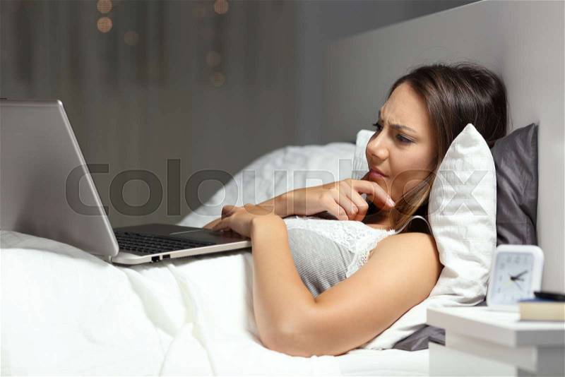 Confused woman using a laptop lying on the bed in the night at home, stock photo