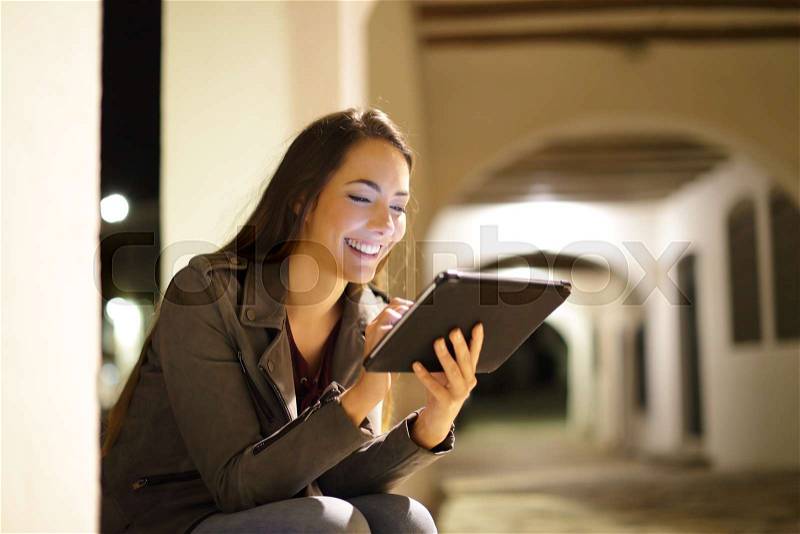 Happy lady browsing tablet content sitting in the night in the street, stock photo