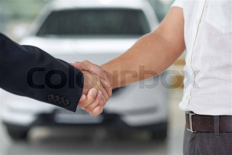 Close up of handshake. Car dealer shaking hands with customer. Men making deal on purchasing automobile in car center. White auto placed in showroom behind. , stock photo