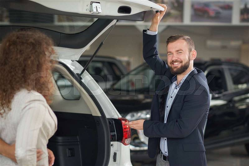 Car dealer showing luggage space to female client. Woman observing details of vehicle, checking quality. Handsome bearded manager smiling, looking at camera, holding ..., stock photo