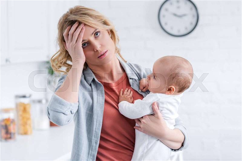Tired mother holding infant baby and standing with hand on forehead at home, stock photo
