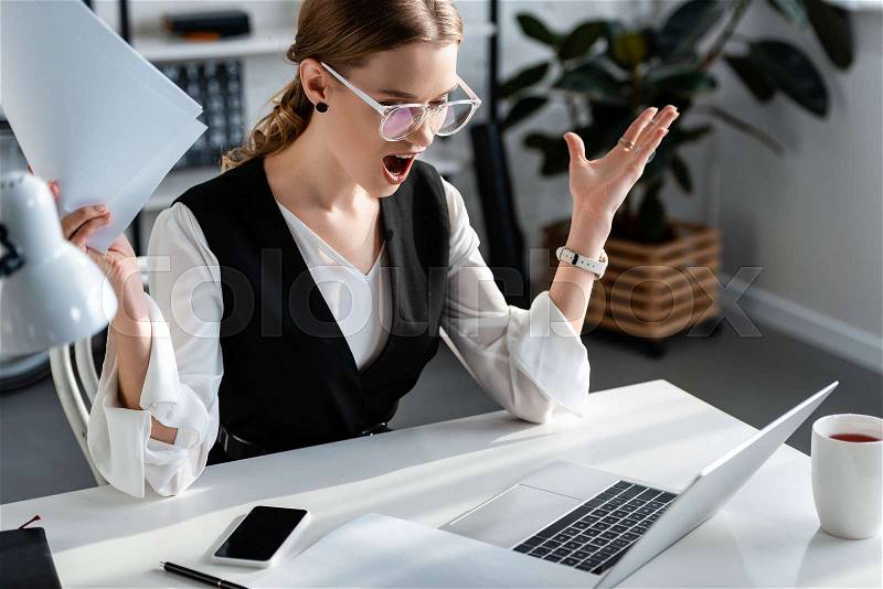 Surprised businesswoman in formal wear sitting at computer desk and gesturing with hands at workplace, stock photo